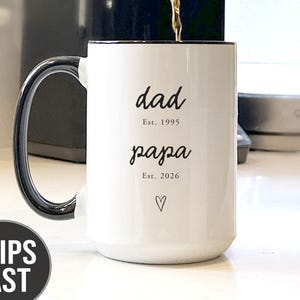 May include: White ceramic mug with black handle and rim, featuring the words "dad" and "papa" in a script font, with the years "Est. 1995" and "Est. 2026" below. A heart symbol is also present. Coffee is being poured into the mug.