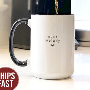 May include: White ceramic mug with a black handle and rim. The mug has the words "aunt melody" and a heart symbol printed on the front. Coffee is being poured into the mug from a coffee maker.
