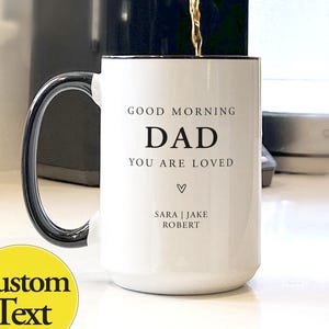 May include: White ceramic mug with a black handle and rim. The mug has the text "GOOD MORNING DAD YOU ARE LOVED" in black, with a heart symbol and the names "SARA | JAKE ROBERT" below. Coffee is being poured into the mug.