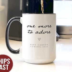 May include: White ceramic mug with a black handle and rim, filled with coffee. The mug has the text "one more to adore" with a heart symbol and "BABY CARSON COMING 2026" printed on it. A coffee maker is pouring coffee into the mug.