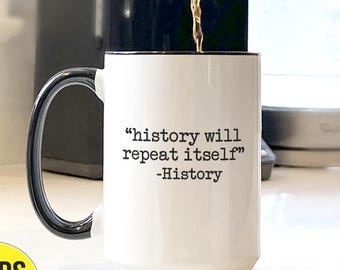 May include: White ceramic mug with a black handle and rim, filled with coffee. The mug features the quote "history will repeat itself" in black text. A yellow sticker with "SHIPS FAST" is visible.