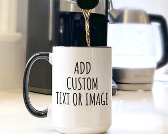 May include: A white ceramic mug with a black handle and interior rim. The front of the mug features the black printed text ADD CUSTOM TEXT OR IMAGE in a simple sans-serif font. The mug is positioned under a coffee machine spout pouring liquid.