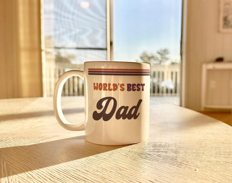 May include: White ceramic mug with the text "WORLD'S BEST Dad" in brown and orange lettering. The mug has a handle and a decorative stripe design in brown, orange, and purple. The mug is sitting on a wooden table.