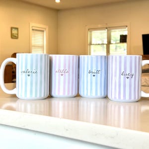 May include: Four ceramic mugs with vertical striped patterns in pastel colors: blue, pink, and lavender. Each mug has a white handle and features a name in cursive with a small heart. The names are "valerie", "stella", "britt", and "lucy".
