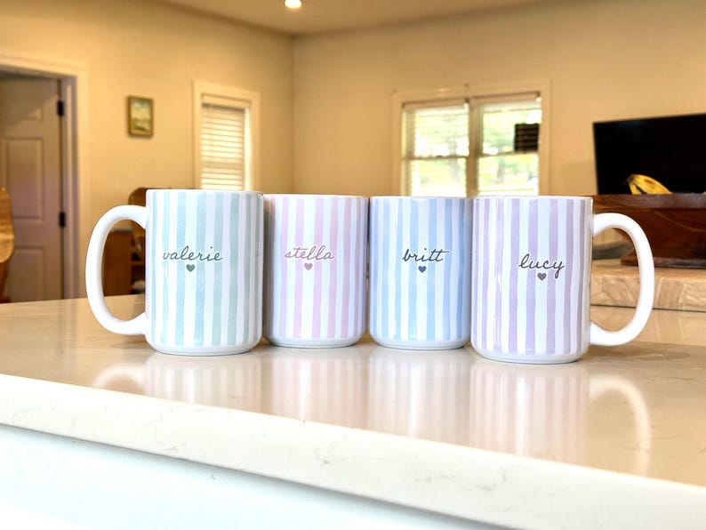 May include: Four ceramic mugs with vertical striped patterns in pastel colors: blue, pink, and lavender. Each mug has a white handle and features a name in cursive with a small heart. The names are "valerie", "stella", "britt", and "lucy".