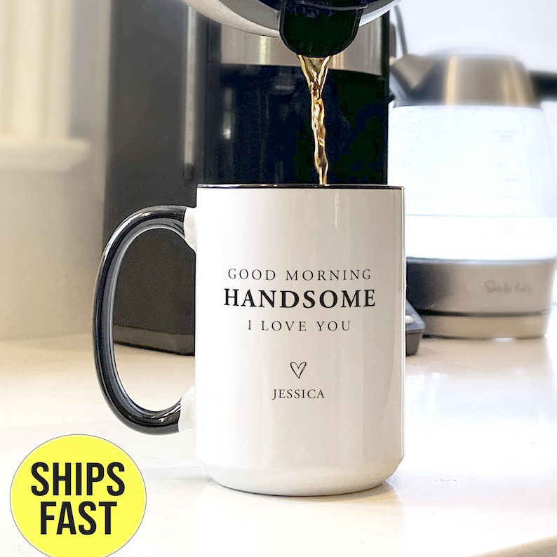 May include: A white ceramic mug with a black handle and rim, featuring the black printed text GOOD MORNING HANDSOME I LOVE YOU, a small heart icon, and the name JESSICA. Liquid is being poured into the mug from a coffee machine above.