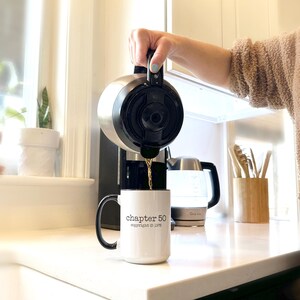 May include: A stainless steel coffee carafe pouring coffee into a white mug with a black handle. The mug has the text "chapter 50 copyright &copy; 1975". A coffee maker and electric kettle are in the background.