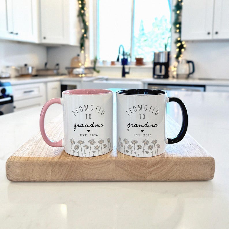 May include: Two white ceramic mugs with the text "PROMOTED to grandma EST. 2026". One mug has a pink handle and interior, the other has a black handle and interior. Both mugs have a floral design at the bottom. They are displayed on a wooden cutting board.