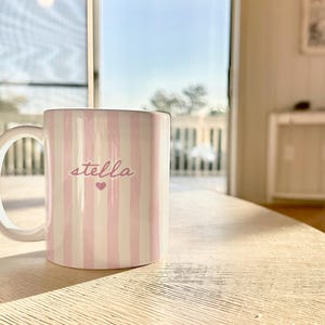 May include: A white ceramic mug with pink vertical stripes and the name "stella" in cursive, with a small heart below the name. The mug is on a light-colored wooden table, with a bright window in the background.