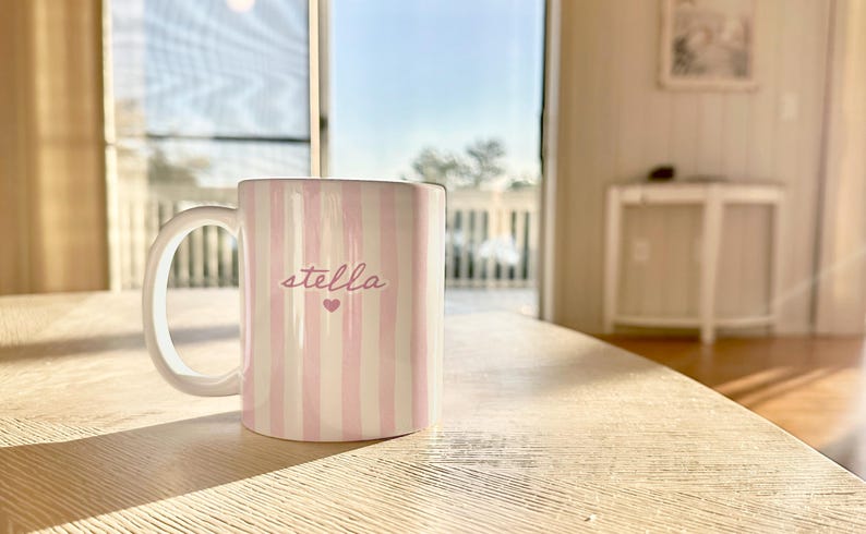 May include: A white ceramic mug with pink vertical stripes and the name "stella" in cursive, with a small heart below the name. The mug is on a light-colored wooden table, with a bright window in the background.