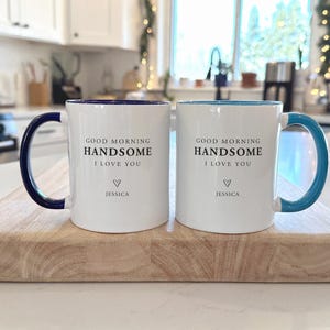 May include: Two white ceramic mugs with coloured handles and interiors. The left mug has a dark blue handle and interior, while the right has a teal one. Both feature the black text: GOOD MORNING HANDSOME I LOVE YOU, with a heart and the name JESSICA.