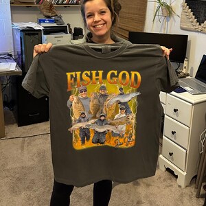 Pode incluir: Uma camiseta cinza escura com uma estampa gr&aacute;fica de um grupo de pessoas pescando. O gr&aacute;fico apresenta o texto "Fish God" e uma assinatura.