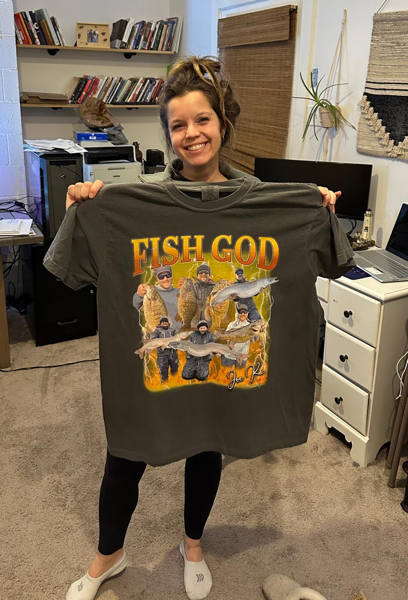 Pode incluir: Uma camiseta cinza escura com uma estampa gr&aacute;fica de um grupo de pessoas pescando. O gr&aacute;fico apresenta o texto "Fish God" e uma assinatura.
