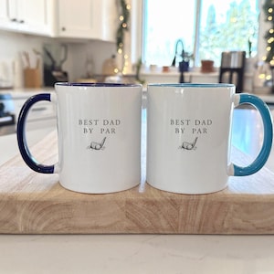 May include: Two white ceramic mugs with the text "BEST DAD BY PAR" and a golf club graphic. One mug has a navy blue handle and rim, the other has a teal handle and rim. They sit on a wooden chopping board.