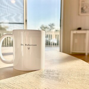 Pu&ograve; includere: Una tazza di ceramica bianca con un manico grande, appoggiata su un tavolo di legno chiaro. La tazza ha il testo "Dr. Robertson" e un simbolo a forma di cuore. Lo sfondo mostra una vista esterna soleggiata.