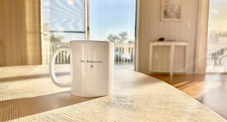 Pu&ograve; includere: Una tazza di ceramica bianca con un manico grande, appoggiata su un tavolo di legno chiaro. La tazza ha il testo "Dr. Robertson" e un simbolo a forma di cuore. Lo sfondo mostra una vista esterna soleggiata.