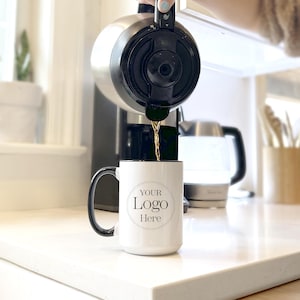 May include: A white ceramic mug with a black rim and handle. The mug has a black circle with the text "YOUR Logo Here" printed on it. Coffee is being poured into the mug from a black coffee maker.