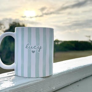 May include: White ceramic mug with vertical light blue stripes. The word "Lucy" is written in cursive, with a small heart below. The mug has a large, curved handle and is set against a blurred outdoor background.