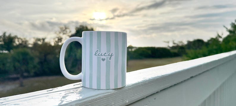 May include: White ceramic mug with vertical light blue stripes. The word "Lucy" is written in cursive, with a small heart below. The mug has a large, curved handle and is set against a blurred outdoor background.