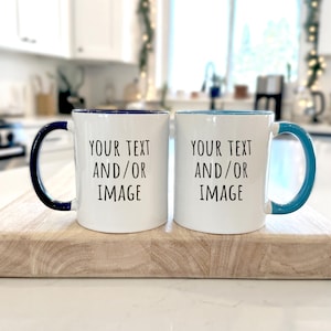 May include: Two white ceramic mugs with blue and teal accents, each with a handle and the text "YOUR TEXT AND/OR IMAGE". The mugs are displayed on a wooden chopping board in a kitchen setting.
