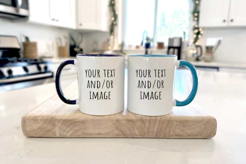 May include: Two white ceramic mugs with blue and teal accents, each with a handle and the text "YOUR TEXT AND/OR IMAGE". The mugs are displayed on a wooden chopping board in a kitchen setting.