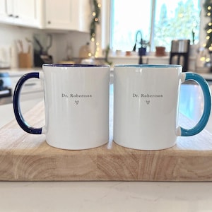 Pu&ograve; includere: Due tazze di ceramica bianche con manici e bordi colorati, appoggiate su un tagliere di legno. Una tazza ha un manico e un bordo blu navy, l'altra ha un manico e un bordo turchese. Entrambe le tazze hanno il testo "Dr. Robertson" e un simbolo a cuore.