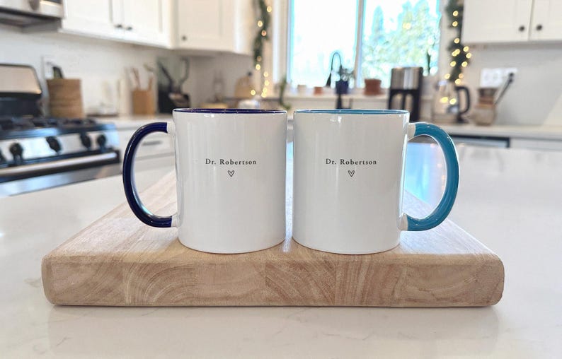 Pu&ograve; includere: Due tazze di ceramica bianche con manici e bordi colorati, appoggiate su un tagliere di legno. Una tazza ha un manico e un bordo blu navy, l'altra ha un manico e un bordo turchese. Entrambe le tazze hanno il testo "Dr. Robertson" e un simbolo a cuore.