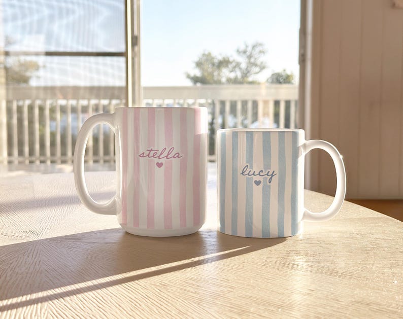 May include: Two ceramic mugs with white handles sit on a wooden table. One mug is pink and white striped with the name "stella" and a heart. The other is blue and white striped with the name "lucy" and a heart.
