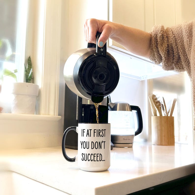 May include: A stainless steel coffee maker pouring coffee into a white mug with black trim. The mug has the text "IF AT FIRST YOU DON'T SUCCEED..." printed on it. The kitchen setting includes a window, a kettle, and a wooden utensil holder.