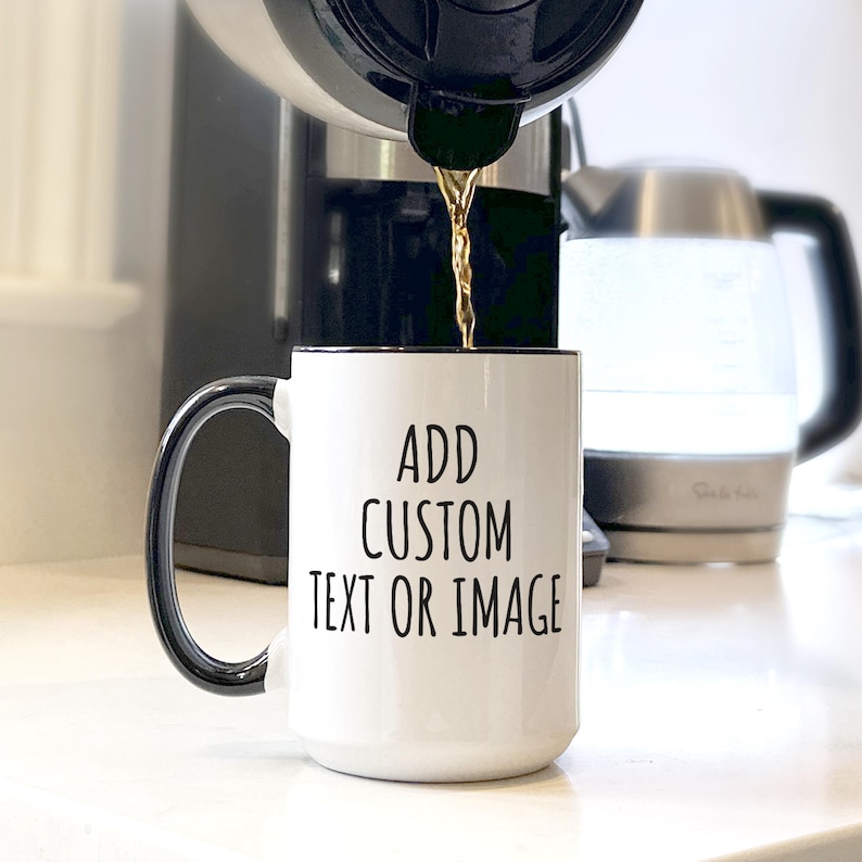 May include: A white ceramic mug with a black handle and rim is being filled with coffee. The mug has the text "ADD CUSTOM TEXT OR IMAGE" in black. A coffee maker and a kettle are in the background.