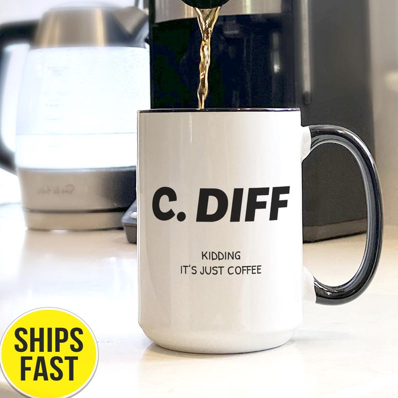May include: White ceramic coffee mug with black rim and handle. The mug has the text "C. DIFF" in bold black letters, with "KIDDING IT'S JUST COFFEE" below. Coffee is being poured into the mug.