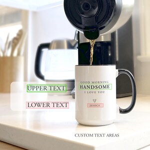 May include: A white ceramic mug with a black handle and rim, featuring the printed text GOOD MORNING HANDSOME I LOVE YOU, a small heart icon, and the name JESSICA. Coffee is being poured into the mug from a coffee maker in the background.