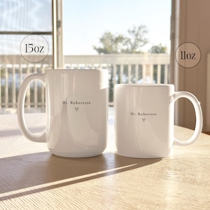 Pu&ograve; includere: Due tazze in ceramica bianca, una da 444 ml e una da 325 ml, sono su una superficie di legno chiaro. Ogni tazza ha un manico e presenta la scritta "Dr. Robertson" con un simbolo a cuore. Le tazze sono davanti a una finestra.
