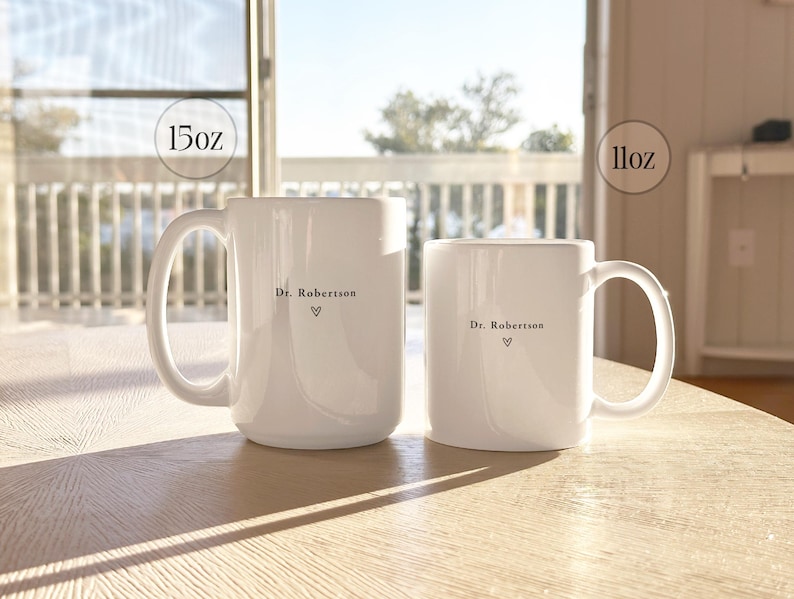 Pu&ograve; includere: Due tazze in ceramica bianca, una da 444 ml e una da 325 ml, sono su una superficie di legno chiaro. Ogni tazza ha un manico e presenta la scritta "Dr. Robertson" con un simbolo a cuore. Le tazze sono davanti a una finestra.