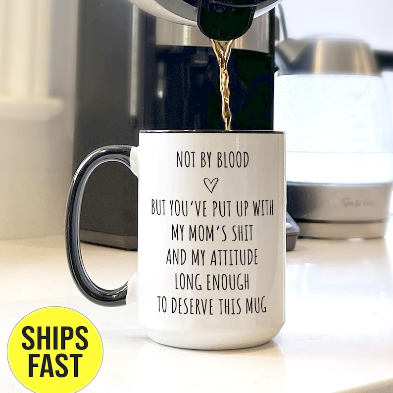 May include: White ceramic mug with a black handle and rim. The mug has the text "NOT BY BLOOD BUT YOU'VE PUT UP WITH MY MOM'S SHIT AND MY ATTITUDE LONG ENOUGH TO DESERVE THIS MUG" printed on it. Coffee is being poured into the mug.