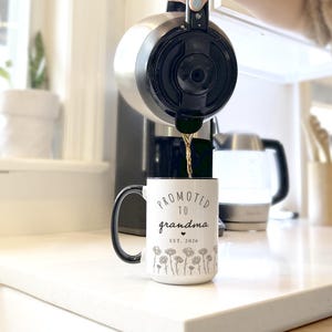 May include: A white ceramic mug with a black handle and rim is being filled with coffee. The mug is printed with the words "Promoted to grandma" and a floral design. The text "EST. 2026" is also printed on the mug.