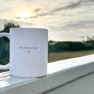 Pu&ograve; includere: Tazza in ceramica bianca con manico curvo. La tazza presenta la scritta "Dr. Robertson" e un simbolo a forma di cuore. La tazza &egrave; appoggiata su una ringhiera bianca con uno sfondo sfocato di alberi e cielo nuvoloso.