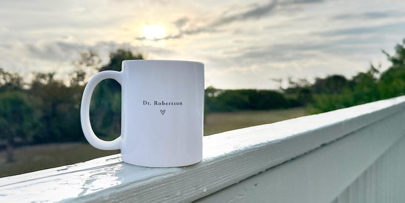 Pu&ograve; includere: Tazza in ceramica bianca con manico curvo. La tazza presenta la scritta "Dr. Robertson" e un simbolo a forma di cuore. La tazza &egrave; appoggiata su una ringhiera bianca con uno sfondo sfocato di alberi e cielo nuvoloso.