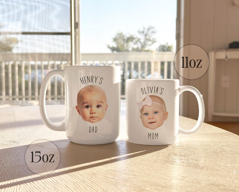 May include: Two white ceramic mugs, one 15oz and one 11oz, with baby photos and text. The larger mug says "Henry's Dad" and the smaller mug says "Olivia's Mom". The mugs are on a light-colored wooden surface.