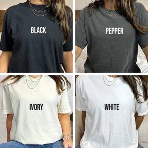 May include: Four short-sleeved t-shirts in black, pepper, ivory, and white. Each shirt has the color name printed in bold, capital letters across the chest. The shirts are displayed on a person.