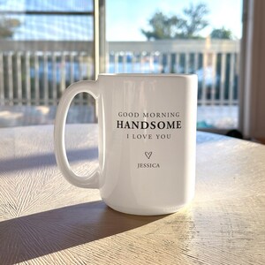 May include: A white ceramic mug with a handle featuring the black printed text GOOD MORNING HANDSOME I LOVE YOU followed by a small heart symbol and the name JESSICA centred on the front surface of the cylindrical drinking vessel.