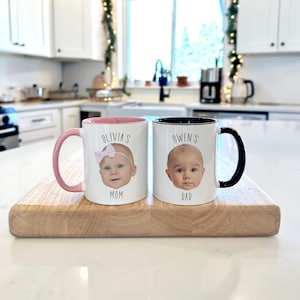 May include: Two white ceramic mugs with baby photos, one with a pink handle and interior, the other with a black handle and interior. The first mug says "Olivia's Mom" and the second says "Owen's Dad". Both mugs are on a wooden cutting board.