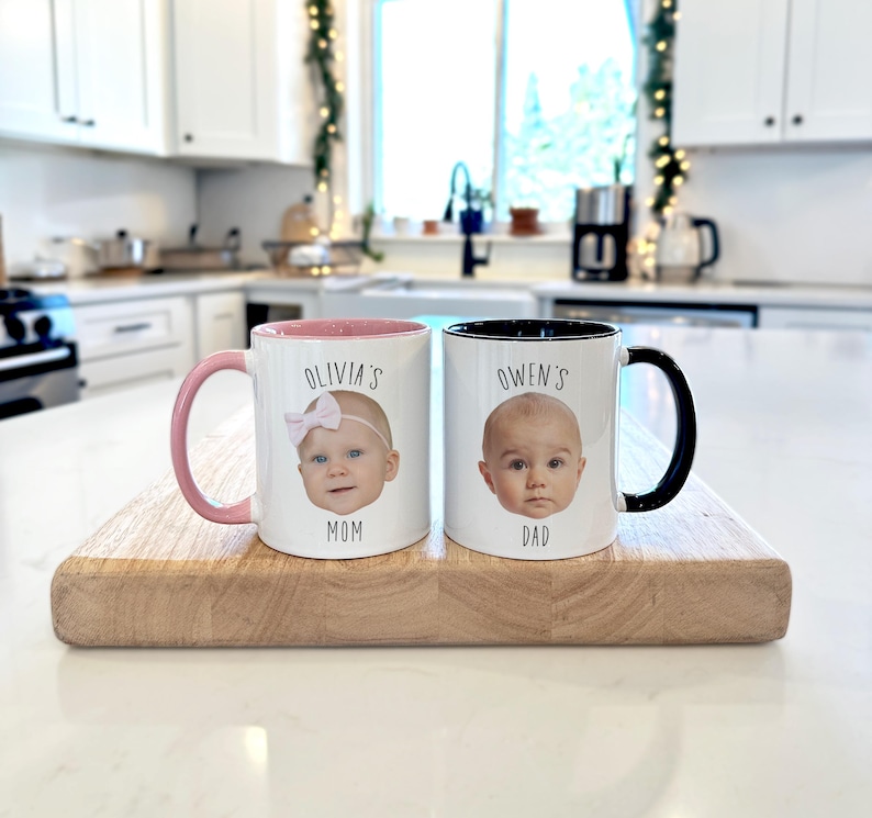May include: Two white ceramic mugs with baby photos, one with a pink handle and interior, the other with a black handle and interior. The first mug says "Olivia's Mom" and the second says "Owen's Dad". Both mugs are on a wooden cutting board.