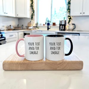 May include: Two white ceramic mugs with black text reading "YOUR TEXT AND/OR IMAGE" are on a wooden board. One mug has a pink interior and handle, the other has a black interior and handle. The background is a bright kitchen setting.