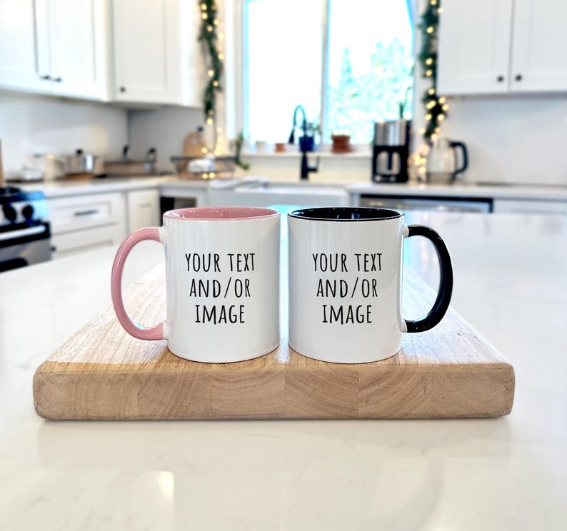 May include: Two white ceramic mugs with black text reading "YOUR TEXT AND/OR IMAGE" are on a wooden board. One mug has a pink interior and handle, the other has a black interior and handle. The background is a bright kitchen setting.