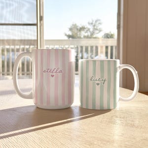 May include: Two ceramic mugs with white handles. One mug is pink and white striped with the name "stella" and a heart. The other mug is green and white striped with the name "lucy" and a heart. Both mugs are on a wooden table.