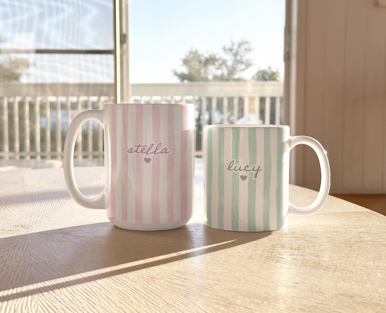 May include: Two ceramic mugs with white handles. One mug is pink and white striped with the name "stella" and a heart. The other mug is green and white striped with the name "lucy" and a heart. Both mugs are on a wooden table.