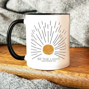 Op de afbeelding: Witte keramische mok met een zwarte handgreep en zwarte rand. De mok heeft een zwarte en gouden zonafbeelding met de tekst "BE THE LIGHT" en "MATTHEW 5:14".