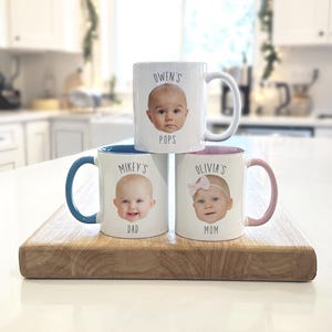 May include: Three white ceramic mugs with baby faces and names printed on them. One mug says "Owen's Pops", another says "Mikey's Dad", and the third says "Olivia's Mom". Each mug has a different colored handle and interior.