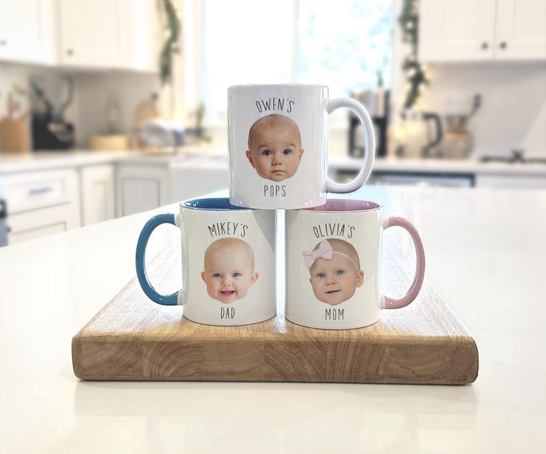May include: Three white ceramic mugs with baby faces and names printed on them. One mug says "Owen's Pops", another says "Mikey's Dad", and the third says "Olivia's Mom". Each mug has a different colored handle and interior.